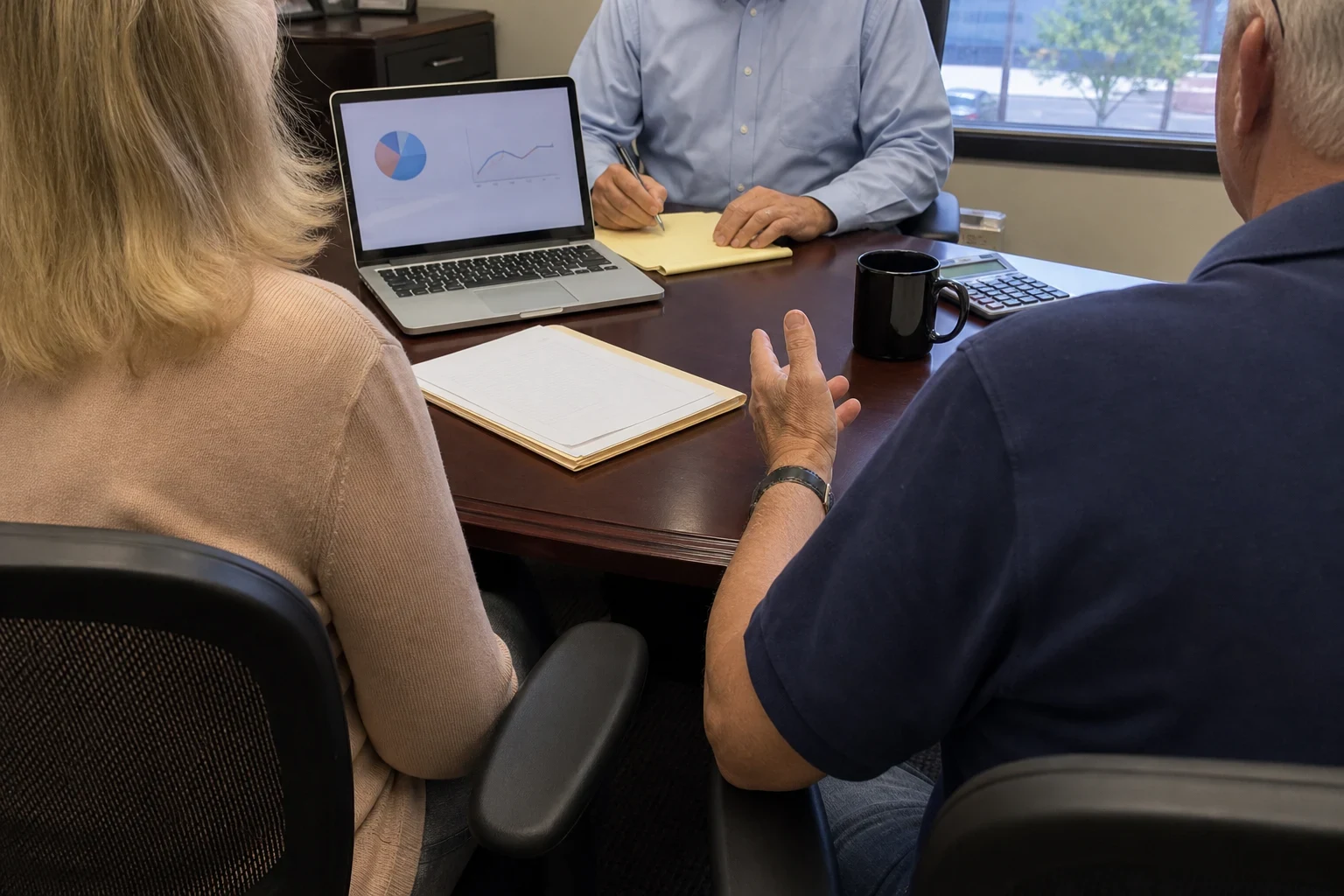 Agent in a light blue button-down taking notes on a legal pad across a conference table from a couple, with a laptop showing charts, a folder, mug and calculator