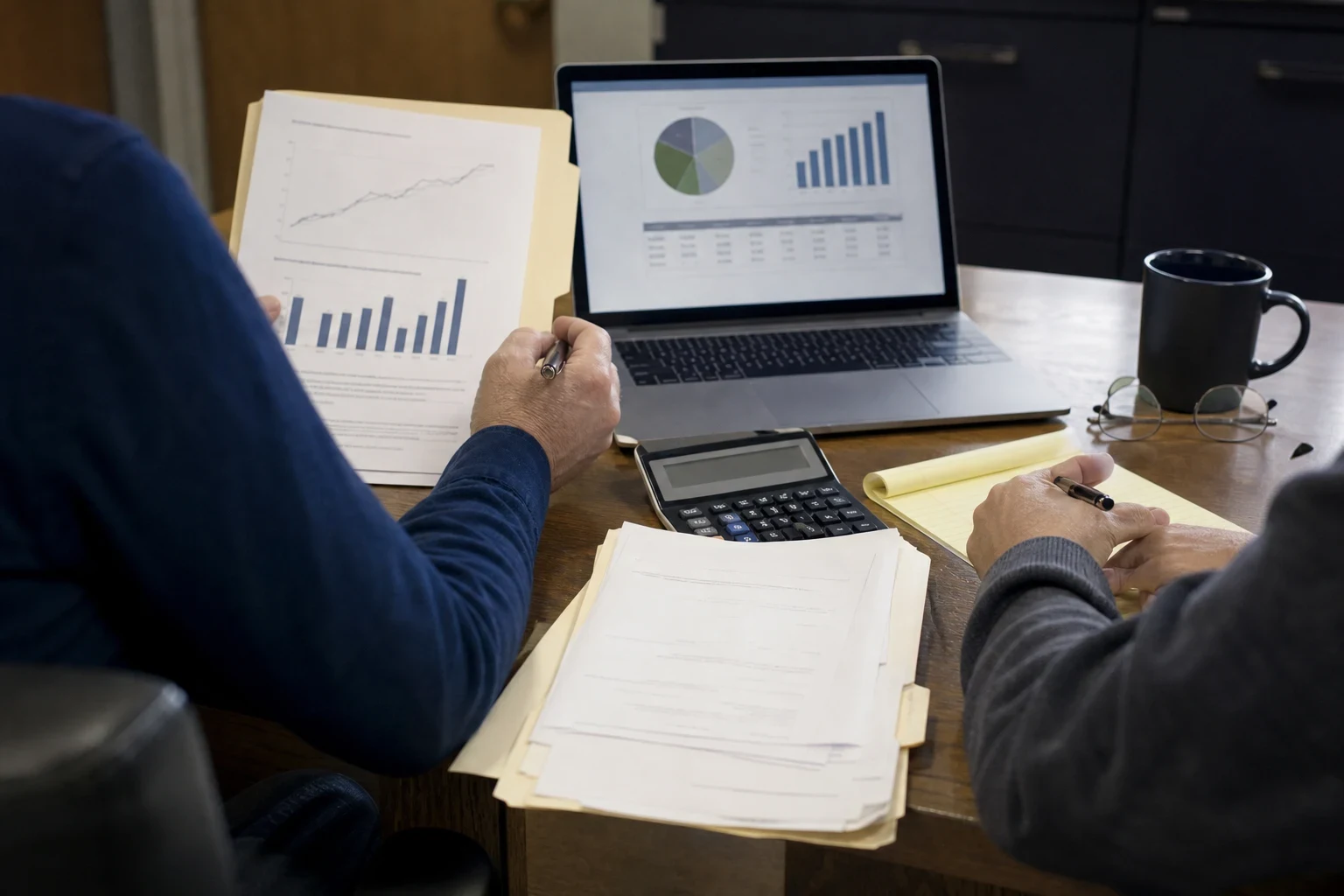 Two people at a wooden office desk reviewing printed charts, a laptop showing pie and bar graphs, a calculator, paperwork and a yellow legal pad